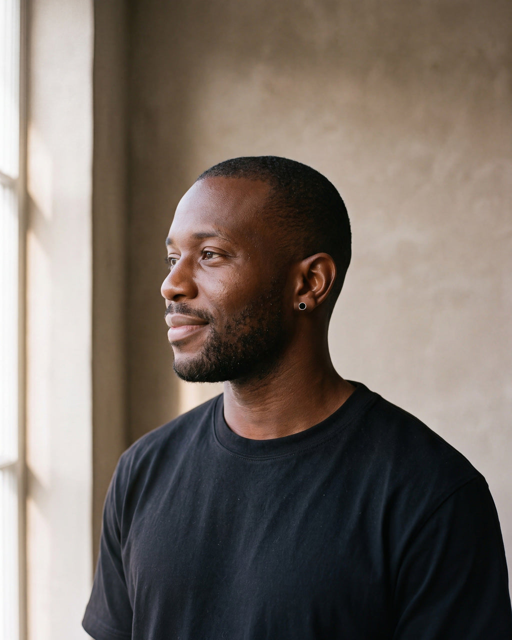 Man in a heavyweight black cotton crew-neck t-shirt by a window wearing the silver Atlas Black Disc Stud Earrings